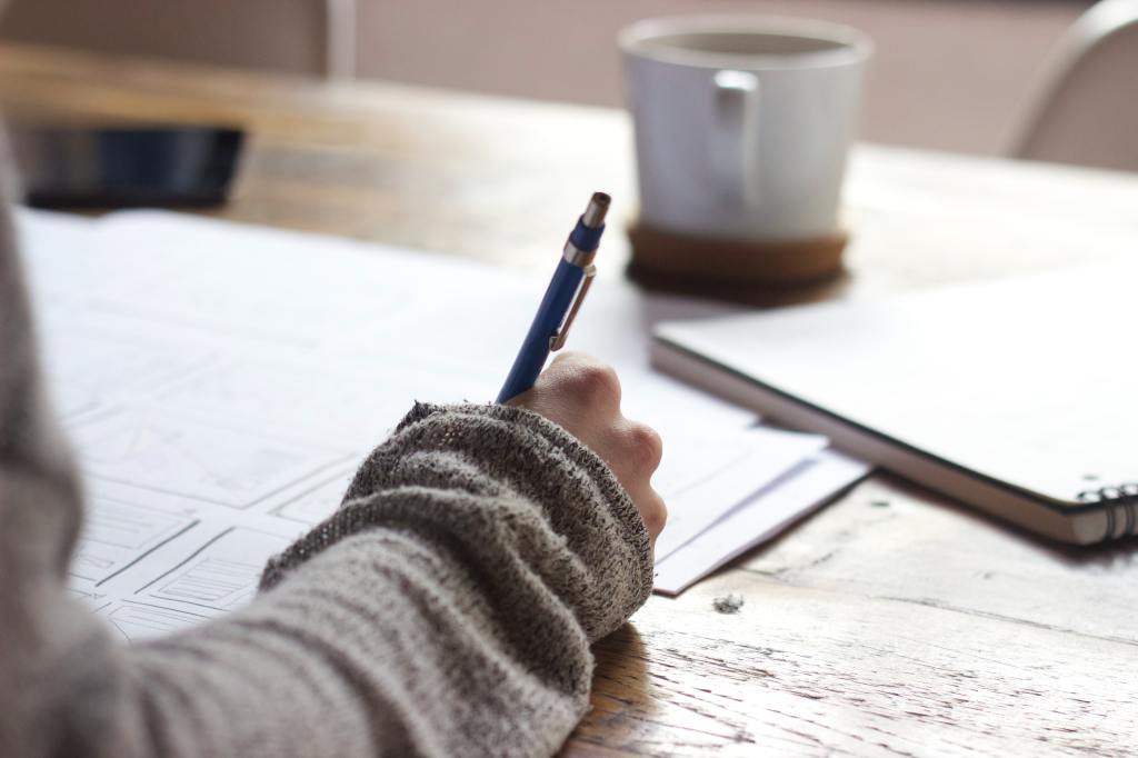 Person writing on brown wooden table near white ceramic mug.