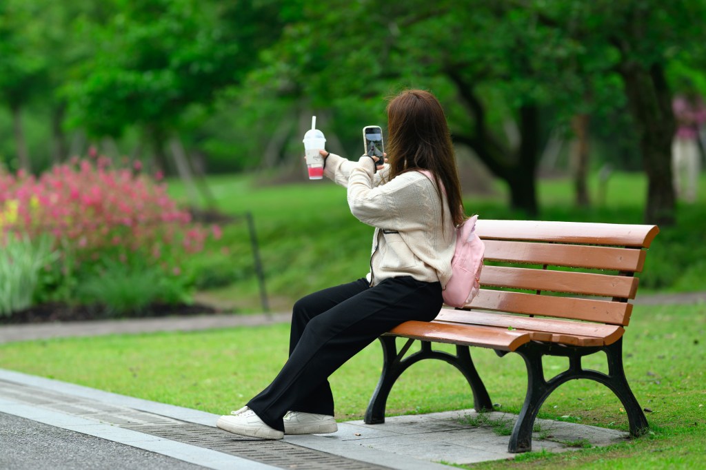 A woman sitting on a bench taking a photo with her cell phone.